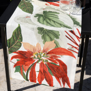 Runner di lino Poinsettia su tavolo scuro, con una grande stella di Natale rossa, foglie verdi e avorio su fondo bianco e bicchieri trasparenti.
