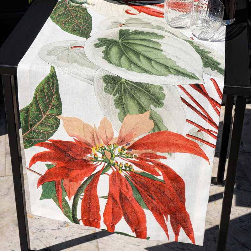 Striscia-da-tavola-Poinsettia-gallery Runner di lino Poinsettia su tavolo scuro, con una grande stella di Natale rossa, foglie verdi e avorio su fondo bianco e bicchieri trasparenti.
