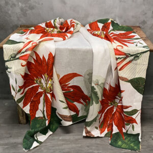 Tovaglia di lino Poinsettia drappeggiata su una panca in legno, con grandi stelle di Natale rosse e foglie verdi su fondo bianco.