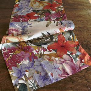 Runner di lino Oleandro appoggiato con morbide pieghe su un tavolo di legno, con fiori corallo, rosa e azzurro-lilla.