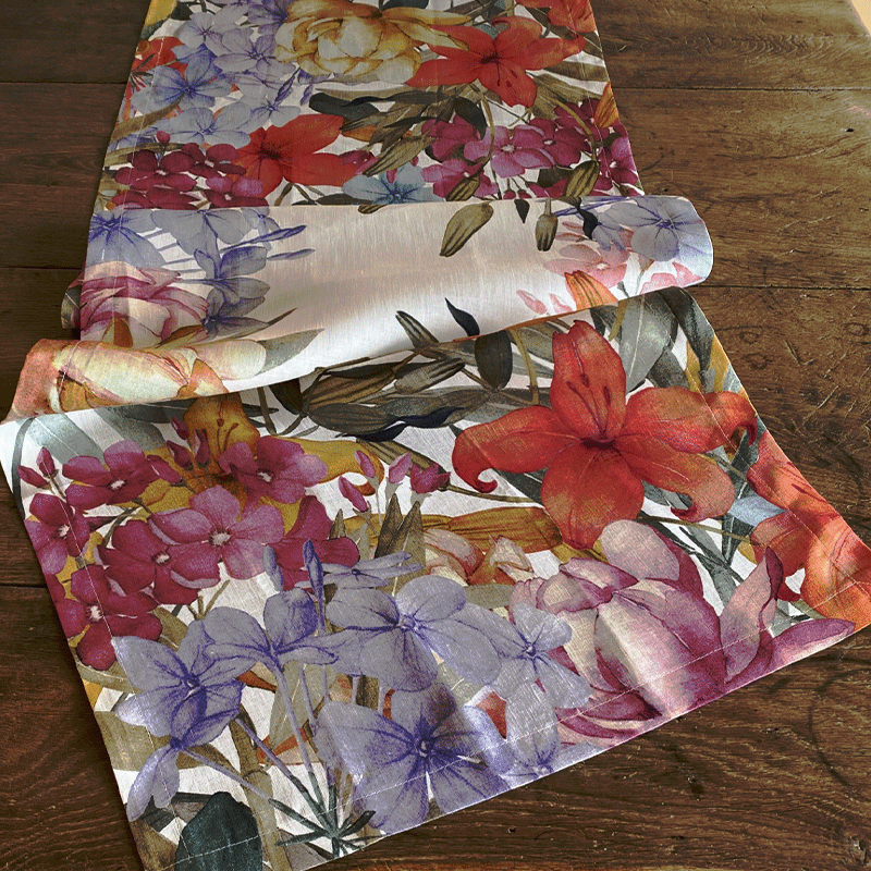 Oleandro Linen Table Runner-la-bottega-di-casa Oleandro linen table runner loosely folded on a wooden table, with coral, pink and lilac-blue flowers.