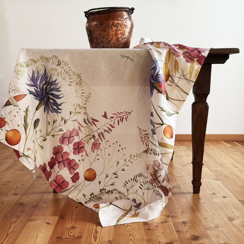 Fiordaliso Linen Tablecloth -la-bottega-di-casa Fiordaliso linen tablecloth by The Napking draped over a wooden table, with blue-violet and pink flowers on a light background.