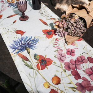 Runner di lino Fiordaliso con fiori blu, rossi e rosa su fondo chiaro, disposto su un tavolo con calice viola e un mazzo di fiori.