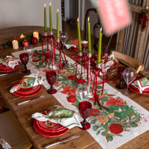 Runner Garland Glee con stelle di Natale rosse, agrumi e foglie verdi su un tavolo di legno con piatti rossi e candele verdi.