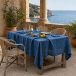 Tovaglia in lino blu cobalto Vintage apparecchiata su una terrazza in pietra tra colonne, con brocca d'argento, bicchieri di cristallo, olive e limoni, vista mare sullo sfondo